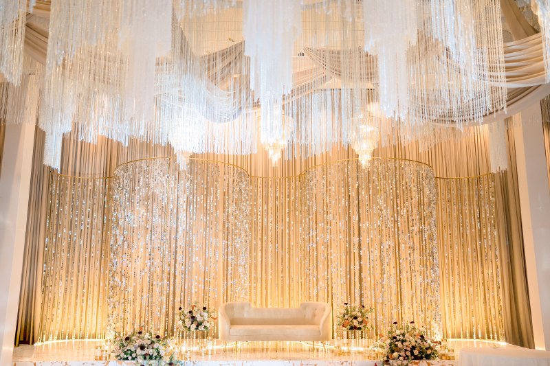 Reception stage with gold crystal curtain backdrop, ivory draping and a white sofa by GPS Decors