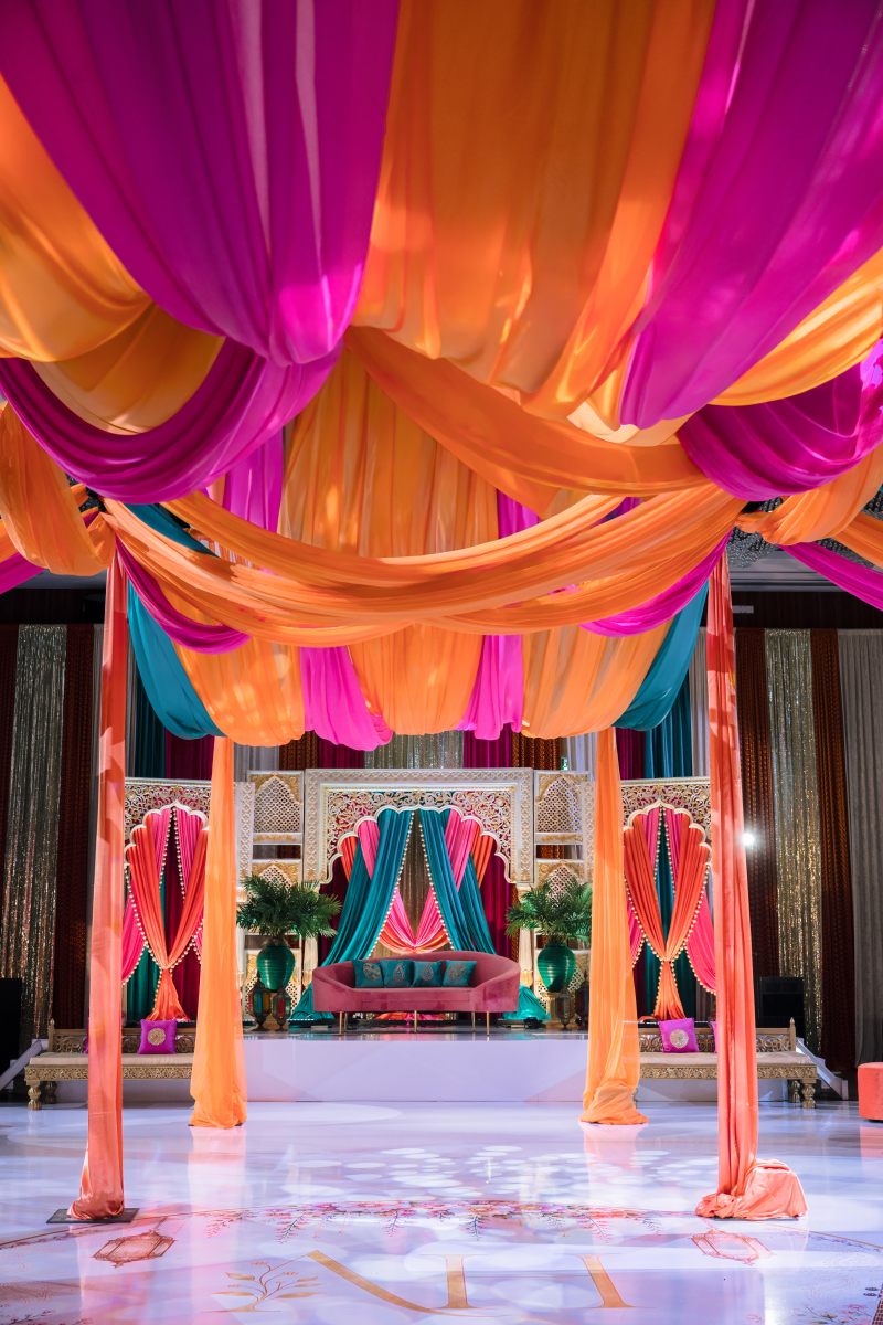 Sangeet ballroom with swagged pink and orange ceiling draping and a jewel-toned stage by GPS Decors
