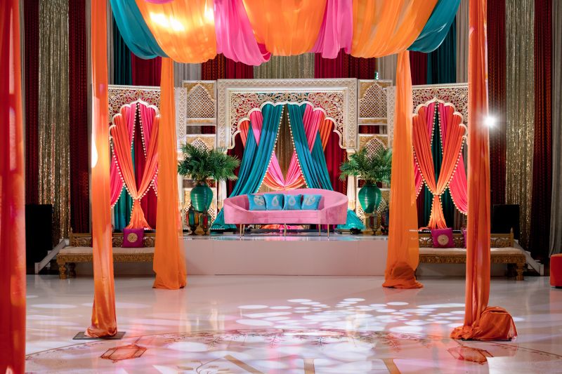 Sangeet stage with carved white panels, teal and pink draping and palms by GPS Decors
