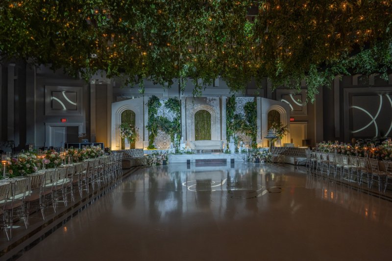 Full room view with a greenery ceiling canopy and fairy lights over long dining tables and a white panel stage with greenery.