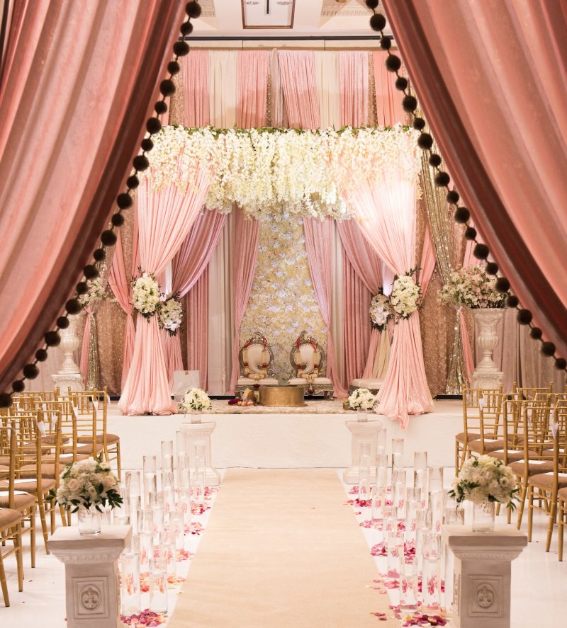 Blush pink draped mandap with white hanging florals, two ornate chairs and an aisle lined with pillars and candles.