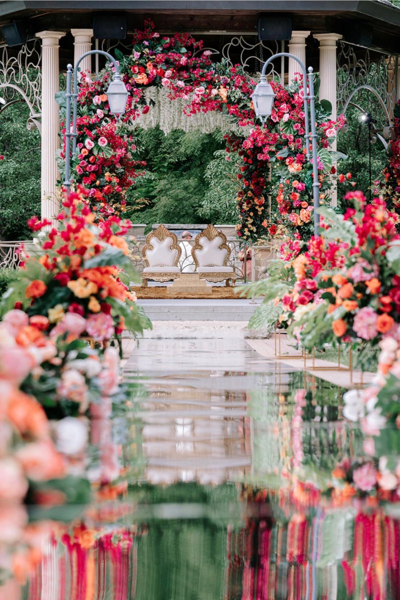 Floral mandap reflected in a mirrored aisle lined with coral, red and pink florals.