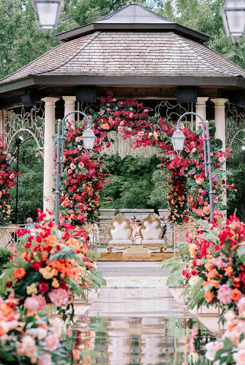 Garden gazebo dressed as a red and pink floral mandap with ornate chairs and a mirrored floral aisle.