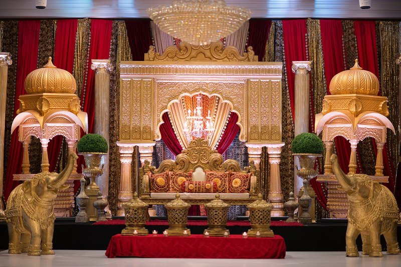 Gold palace-style mandap stage with domed pavilions, gold elephant statues, red drapes and an ornate throne seat.
