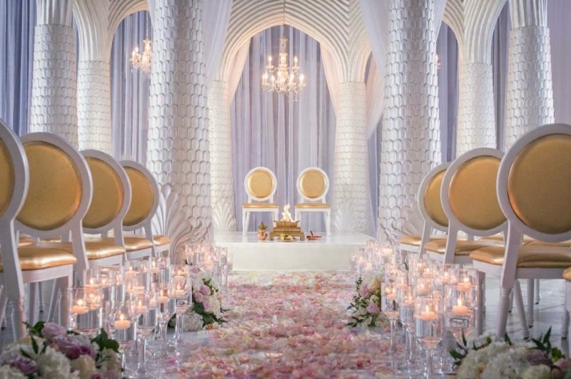 White sculpted palm-style mandap with gold chairs and havan setup at the end of a candlelit petal aisle.