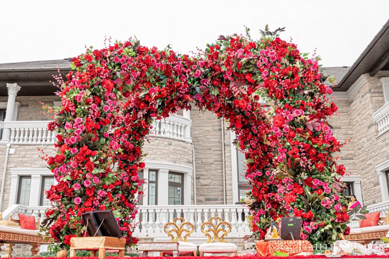 Outdoor mandap built from two arches of red and pink roses with ornate gold chairs and ceremony setup in front of a stone estate.