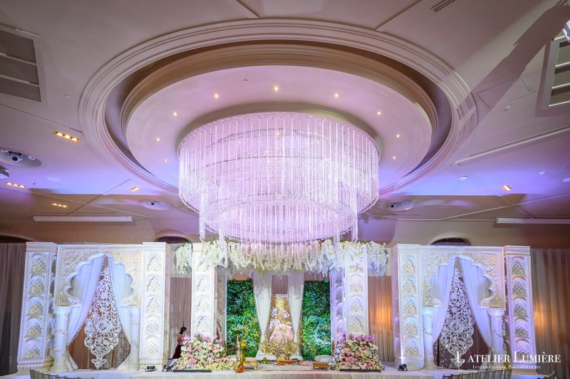 White carved jali mandap with a greenery backdrop beneath a large crystal chandelier in a ballroom.