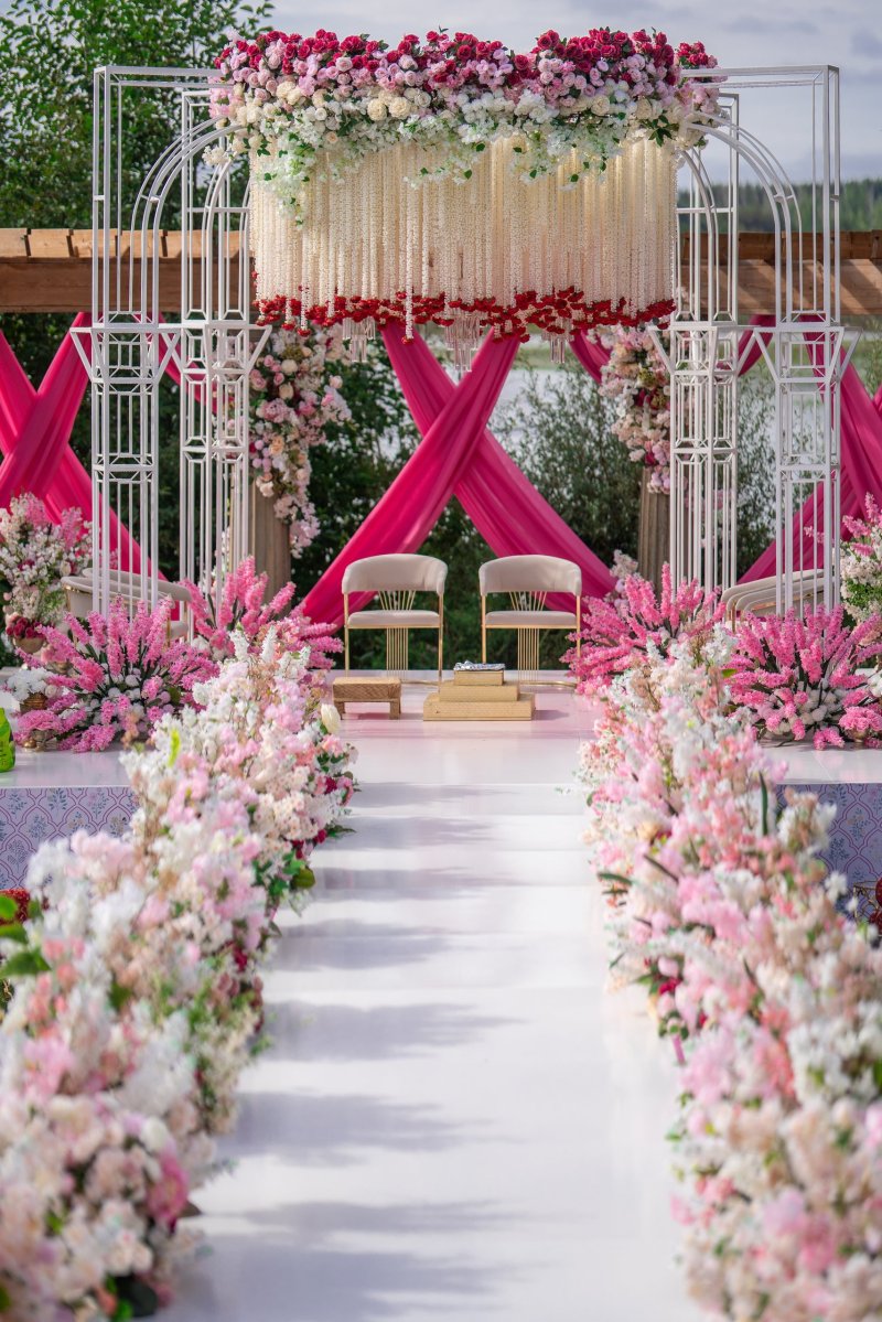 White lattice mandap with crossed pink drapes, hanging white florals and a pink floral aisle under an outdoor pergola.