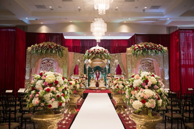 Ornate mandap with red draping, gold urns of white and red roses and a white aisle at Copper Creek Golf Club by GPS Decors