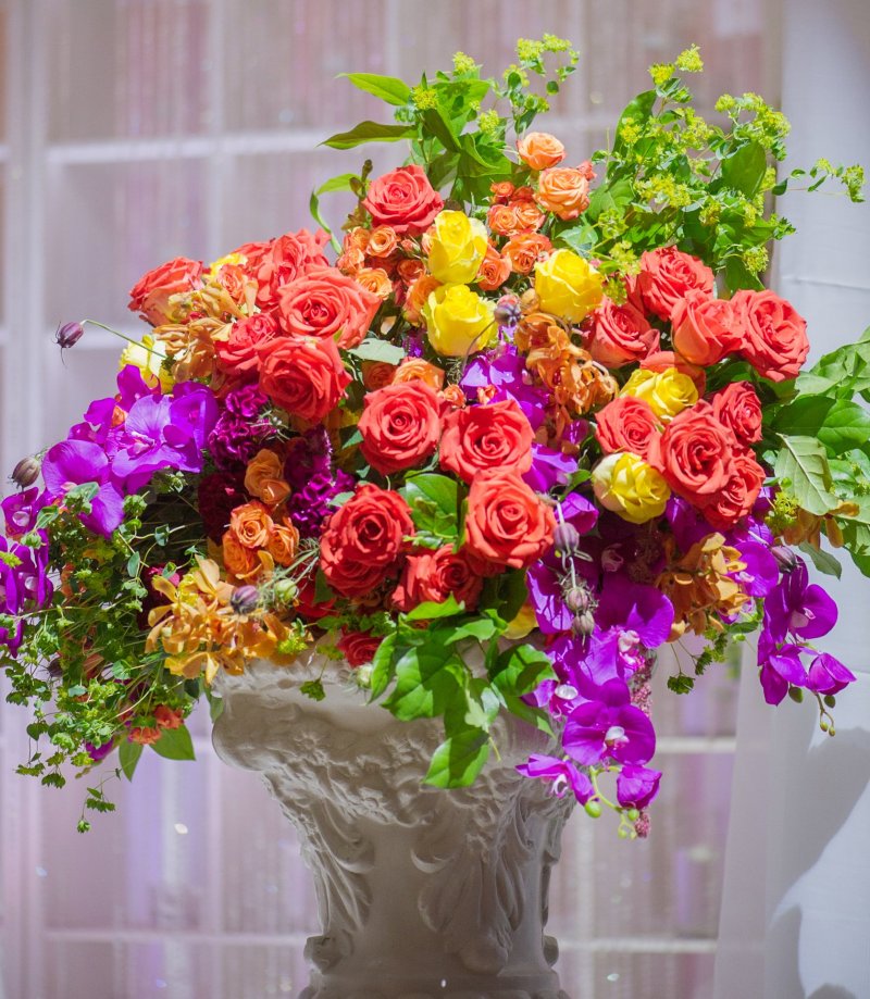Bright floral arrangement of coral roses, yellow roses and purple orchids in a white urn at a mehndi party by GPS Decors