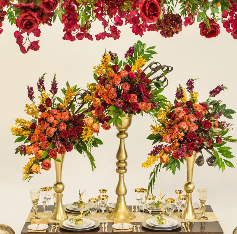 Three gold vase centrepieces of orange and red roses with orchids beneath a hanging floral installation by GPS Decors