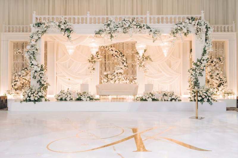 White floral arch stage with greenery and a gold monogram dance floor at Queens Manor by GPS Decors
