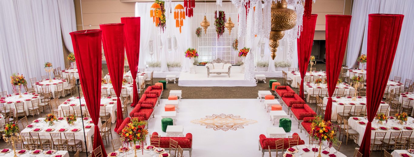 Sangeet hall with red draped columns, red and orange floor seating and a white stage by GPS Decors
