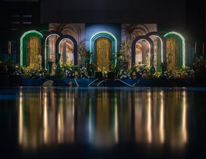Dark stage with neon-outlined arches, palm-print panels, gold curtains and white florals reflected in a glossy floor.