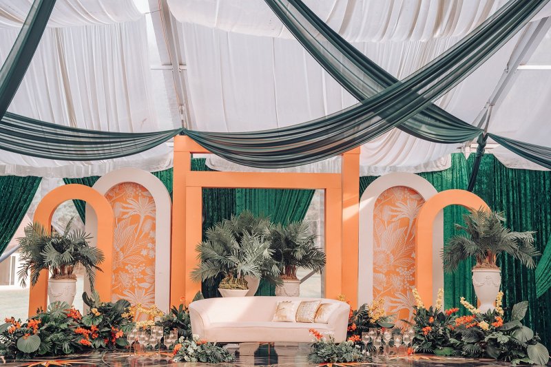 Tented stage with orange arch panels in a tropical print, palms, orange florals, a white sofa and green ceiling drapes.