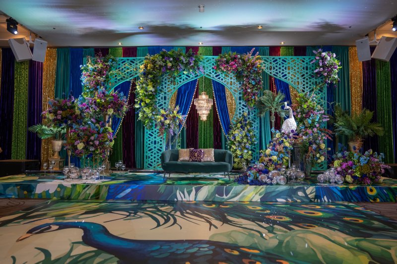 Peacock-themed stage with teal jali arches, jewel-tone drapes, colourful florals, peacock figures and a peacock-print dance floor.