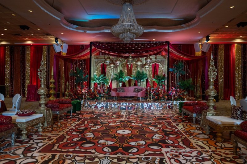 Ballroom with red and gold drapes, a canopy stage with lanterns and palms, a pink sofa, gold low seating and a colourful printed dance floor.