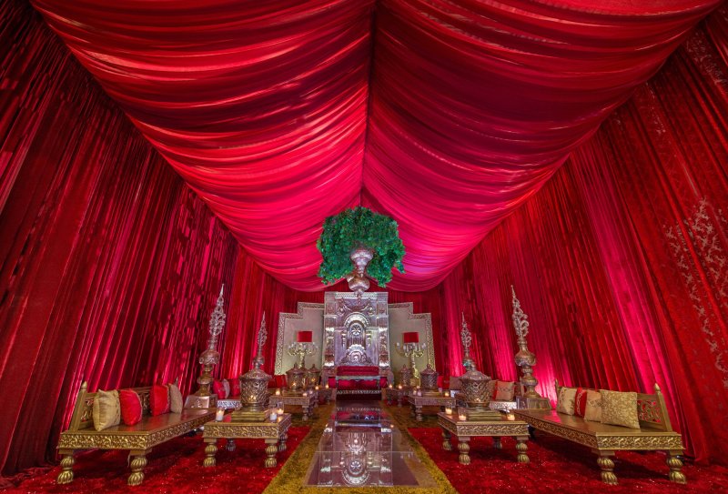 Red velvet draped tent ceiling over a silver ornate throne backdrop, gold low seating with cushions and a mirrored aisle.