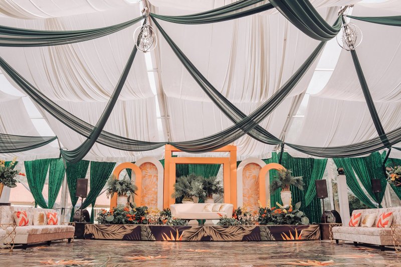 Wide view of a tent with green and white ceiling drapes, an orange arch stage with palms and lounge seating on either side.