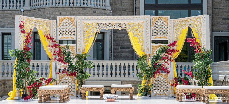 Outdoor white and gold carved jali backdrop with yellow drapes, bougainvillea and low carved wooden seating.