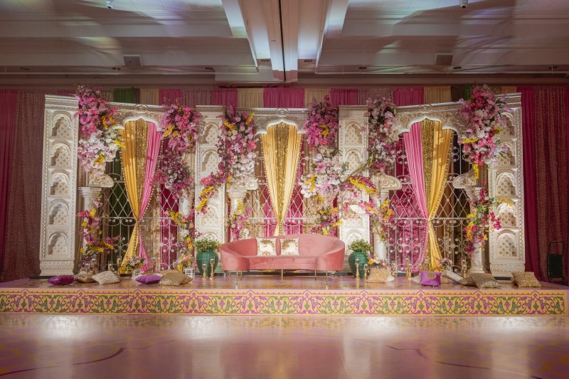 White carved jali stage with pink and yellow drapes, pink and purple orchid florals, a pink velvet sofa and floor cushions.