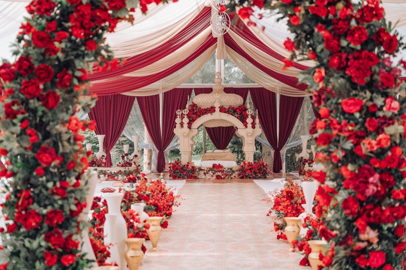 Red rose arch and burgundy draped tent leading to a carved palki at MG Manor by GPS Decors