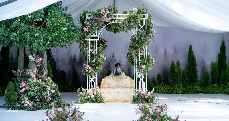 Anand Karaj palki of white lattice wrapped in pink and green florals beside trees, in front of a misty forest backdrop.
