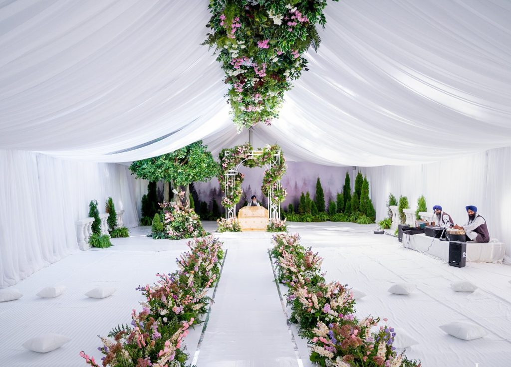 Wide view of a white tented Anand Karaj space with a floral-lined aisle, hanging floral installation and lattice palki.