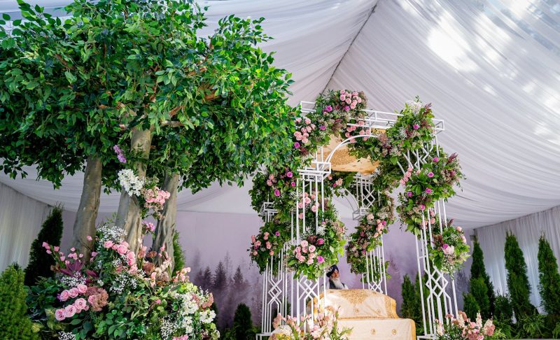 White lattice palki with pink florals framed by tall greenery trees in a tented ceremony space.