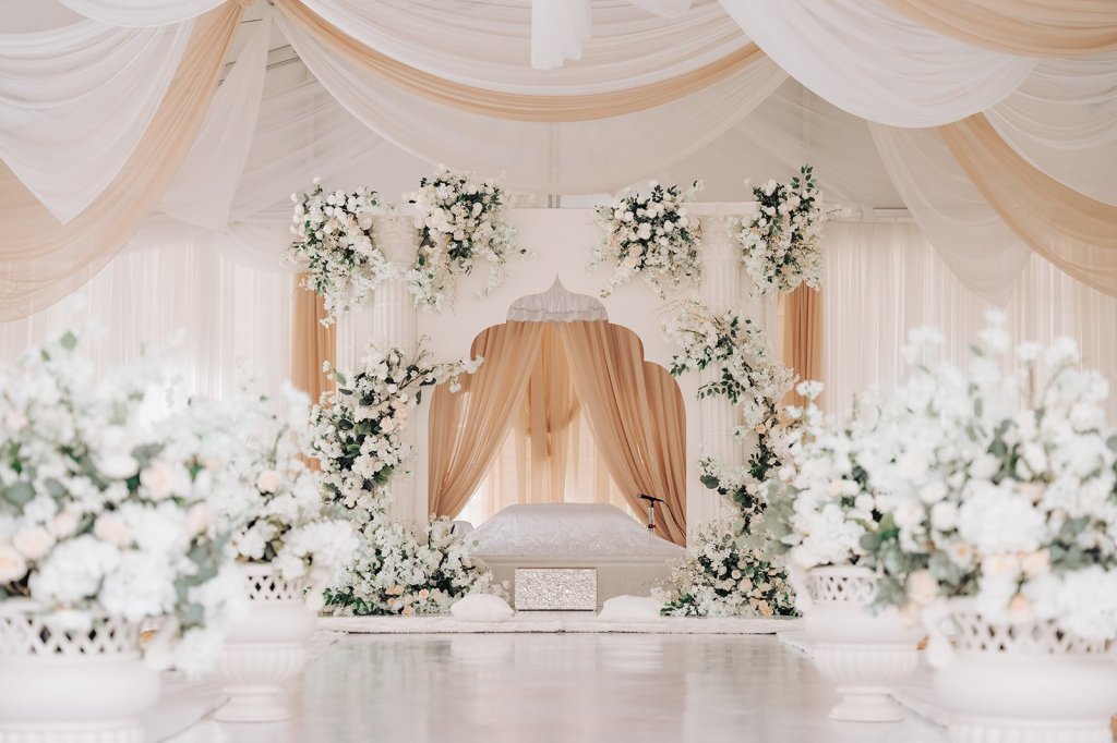 White and peach arched palki surrounded by white floral arrangements under draped tent ceiling.