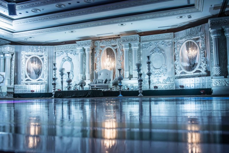 Ballroom lined with white carved panels and mirrors, candelabras and a throne chair above a reflective floor.