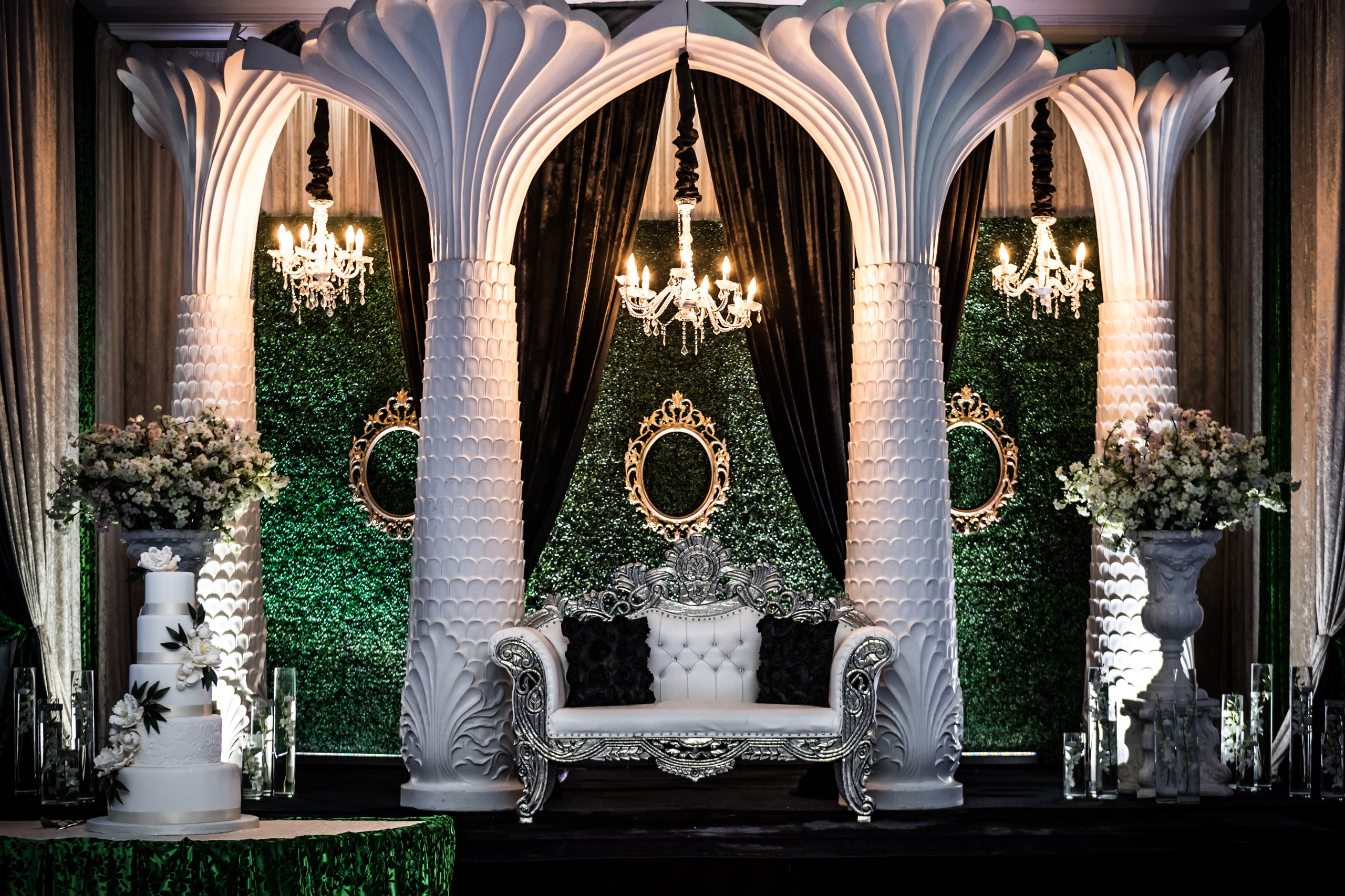 Stage with white palm-style columns, a greenery wall with gold frames, a silver tufted throne loveseat and tiered cake.