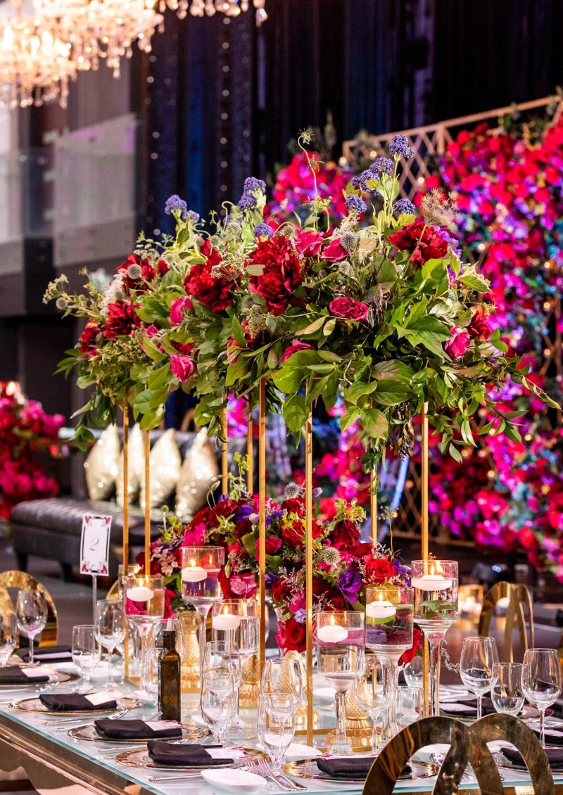 Tall red and pink floral centrepiece on a gold stand with candles in front of a floral wall by GPS Decors