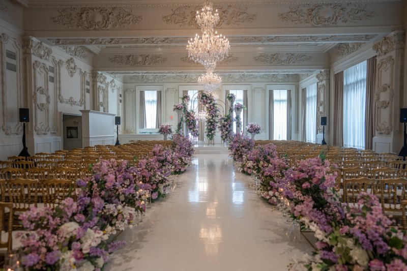 Ceremony in an ornate white ballroom with an aisle lined by lavender and purple floral arrangements leading to floral arches under a crystal chandelier.