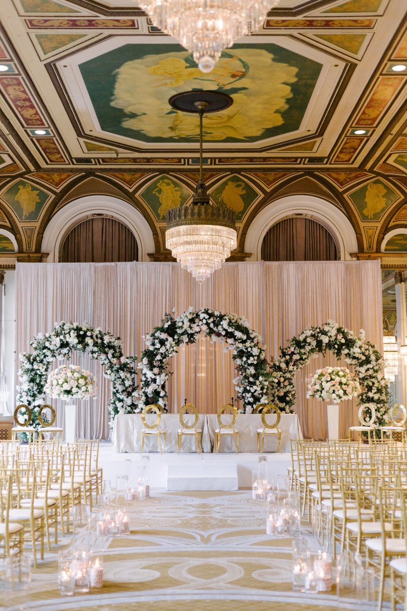 Ceremony in an ornate ballroom with three white floral arches, gold chairs and a candlelit aisle.