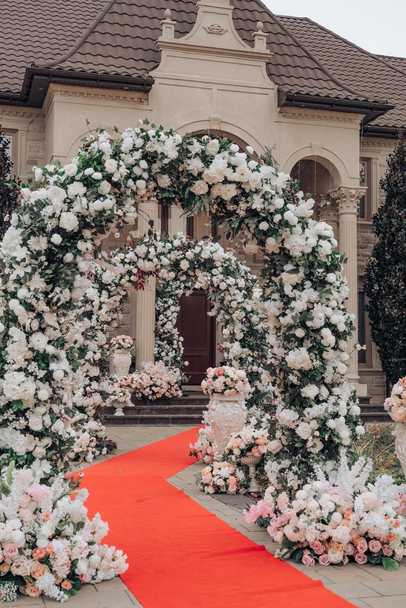 White and pink floral arch over a red carpet aisle for a wedding at home by GPS Decors