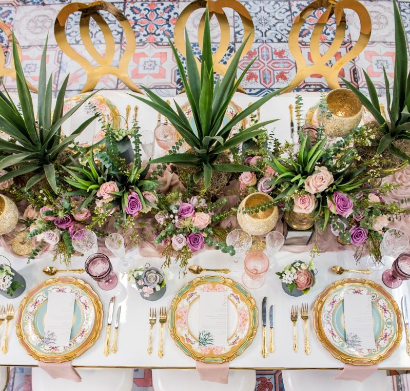 Overhead view of a long table with agave, pink and purple roses, gold chargers and gold chair backs on a patterned tile floor.