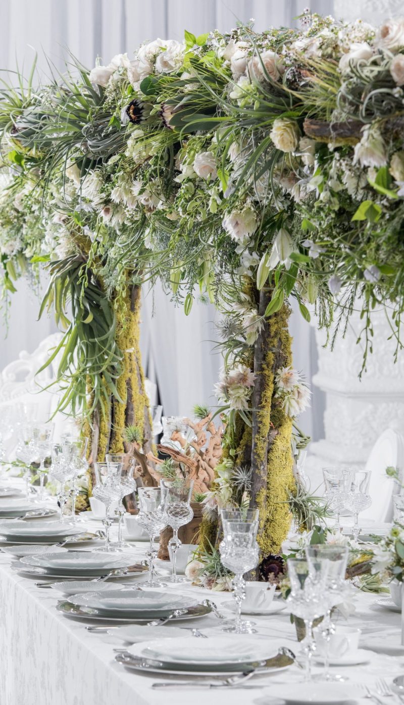 Tall greenery and white floral canopy rising above a long white-linen dining table.