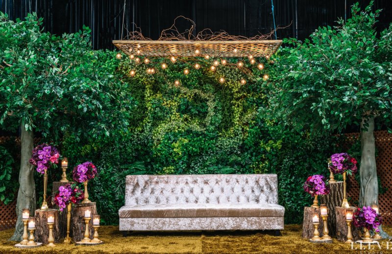 Greenery wall backdrop with monogram, trees, a velvet tufted sofa, purple orchids and candles on tree stumps.