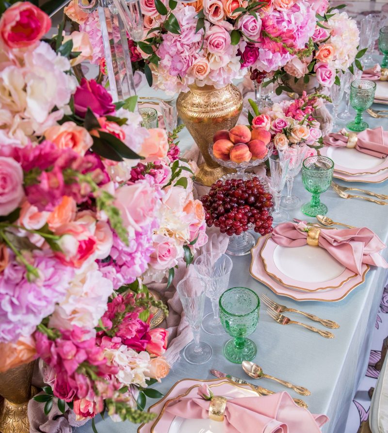 Close-up of pink floral centerpiece with fruit, green goblets and blush napkin place settings.