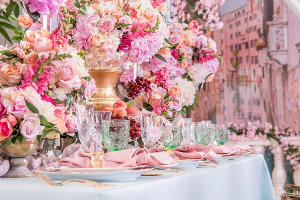 Tablescape with pink rose arrangements in gold vases, green goblets and blush napkins on pale blue linen.