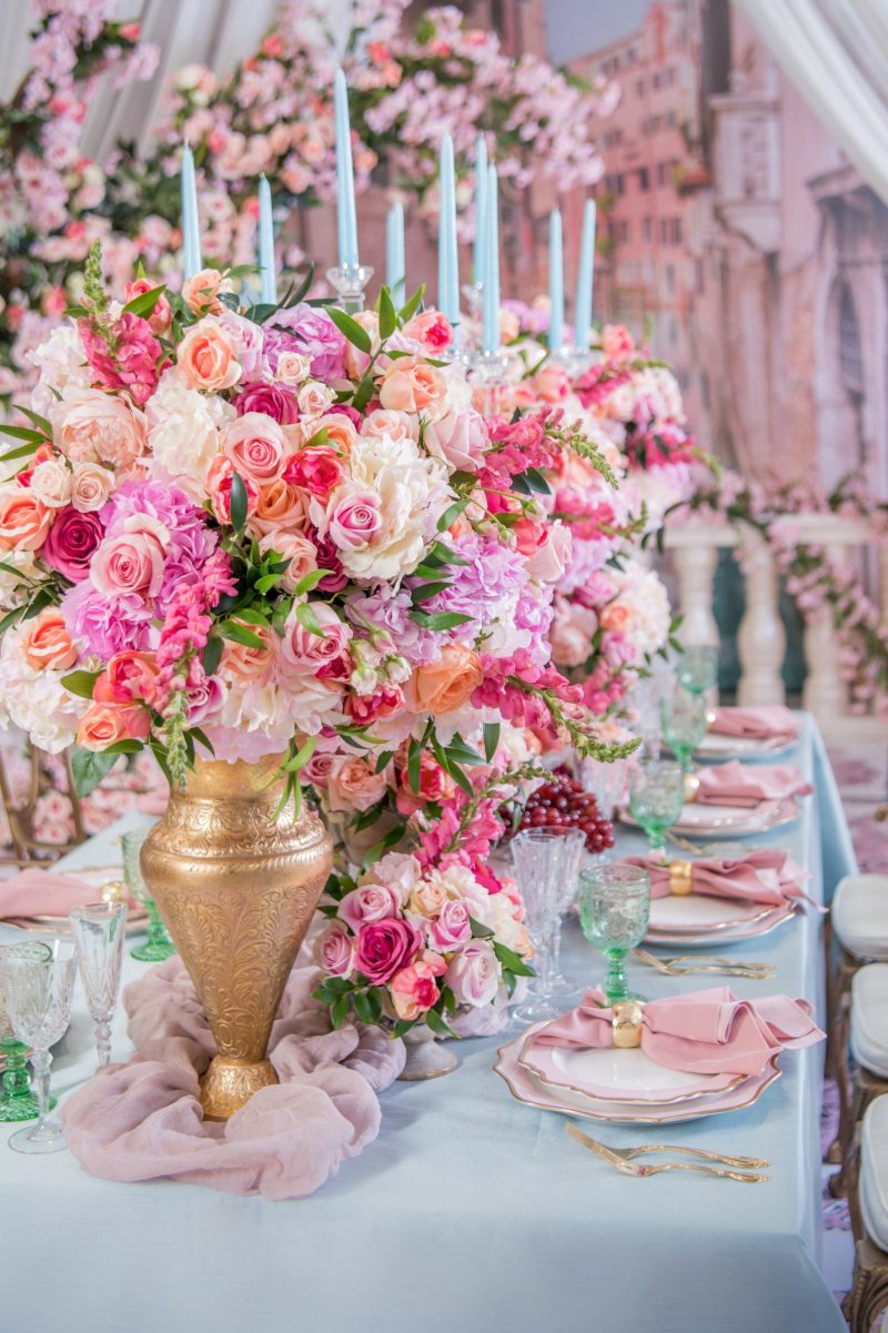 Tall gold vase of pink and peach flowers with pale blue taper candles at the end of a dining table.