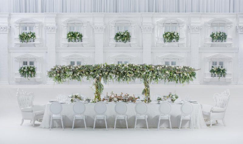 All-white styled long dining table with a greenery canopy, white chairs and a white architectural backdrop.