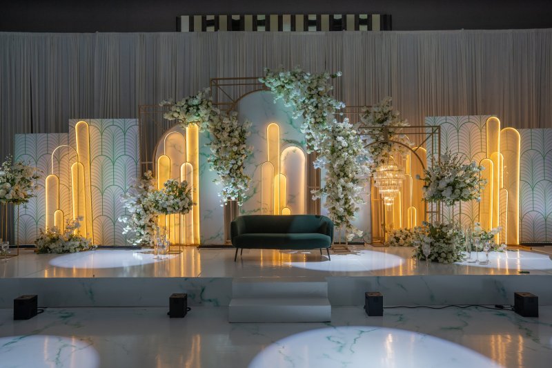 Art deco stage with gold-lit arch panels, white florals and a green velvet sofa.