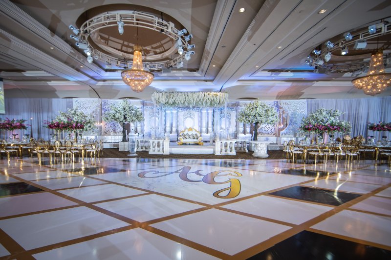 Wide view of a ballroom reception with a white stage, blossom trees, gold chairs and a monogram dance floor under chandeliers.