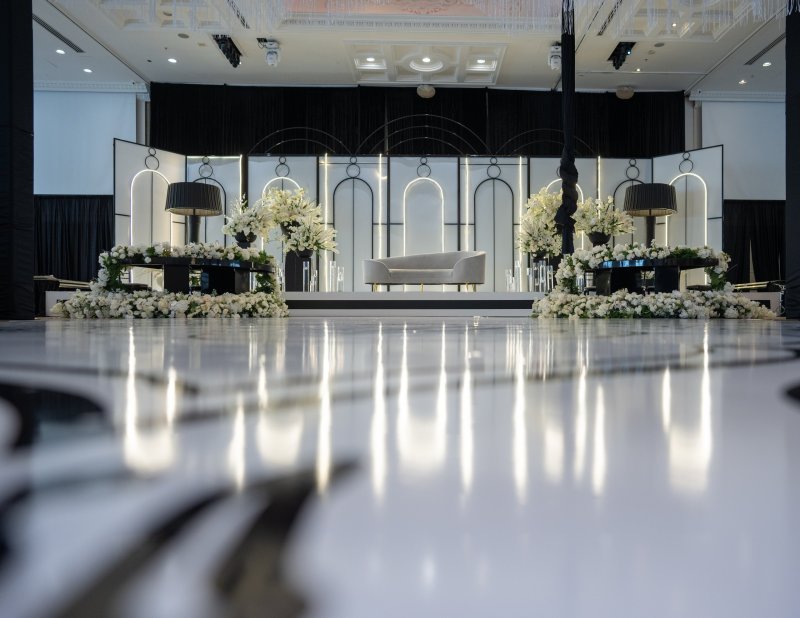 Black and white stage with illuminated arch outlines, white florals, lamps and a curved sofa above a glossy dance floor.