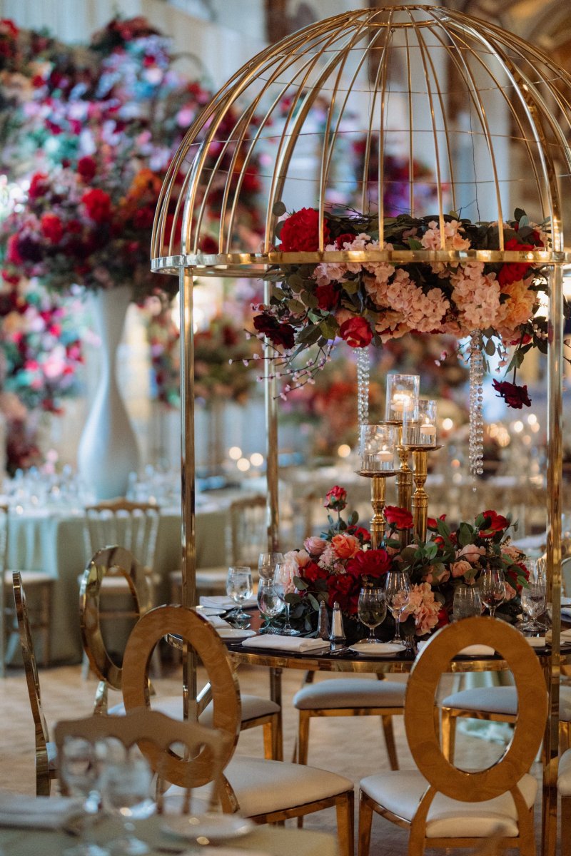Gold birdcage centerpiece filled with red and pink flowers above a table with gold ring-back chairs.