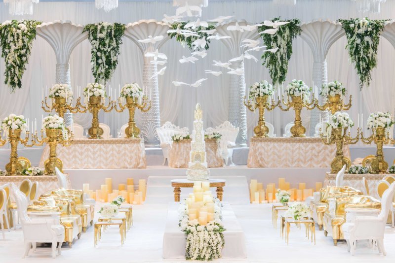 White palm-style columns with hanging greenery and white doves, gold candelabras with white roses, a tiered cake and gold lounge seating.