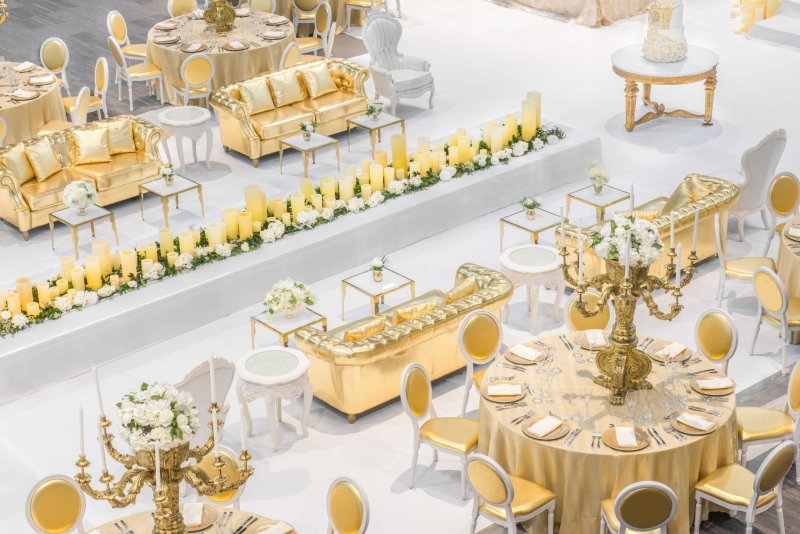 Overhead view of gold tufted sofas, gold-linen round tables and a long candle runner on a white floor.