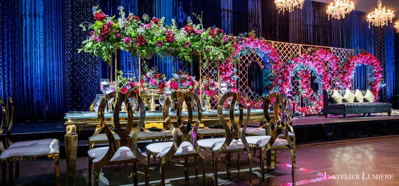 Long head table with gold ring-back chairs on a stage, set against floral ring backdrops of pink and red roses and blue velvet drapes.